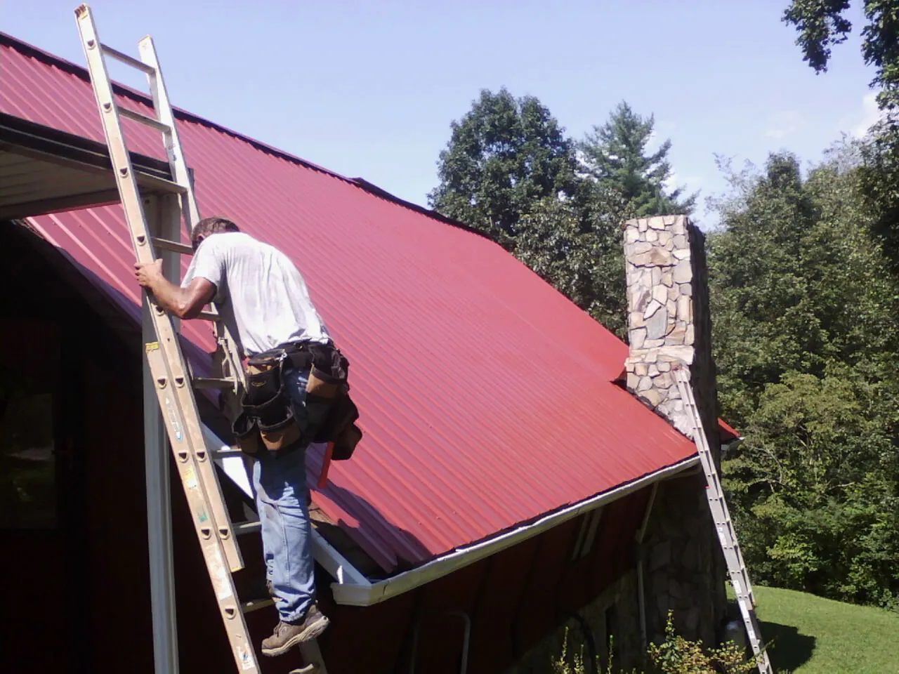 Roofer on a ladder beside a red metal roof and stone chimney — Soffit & Fascia Repair by Roofing Brothers in Hartwell